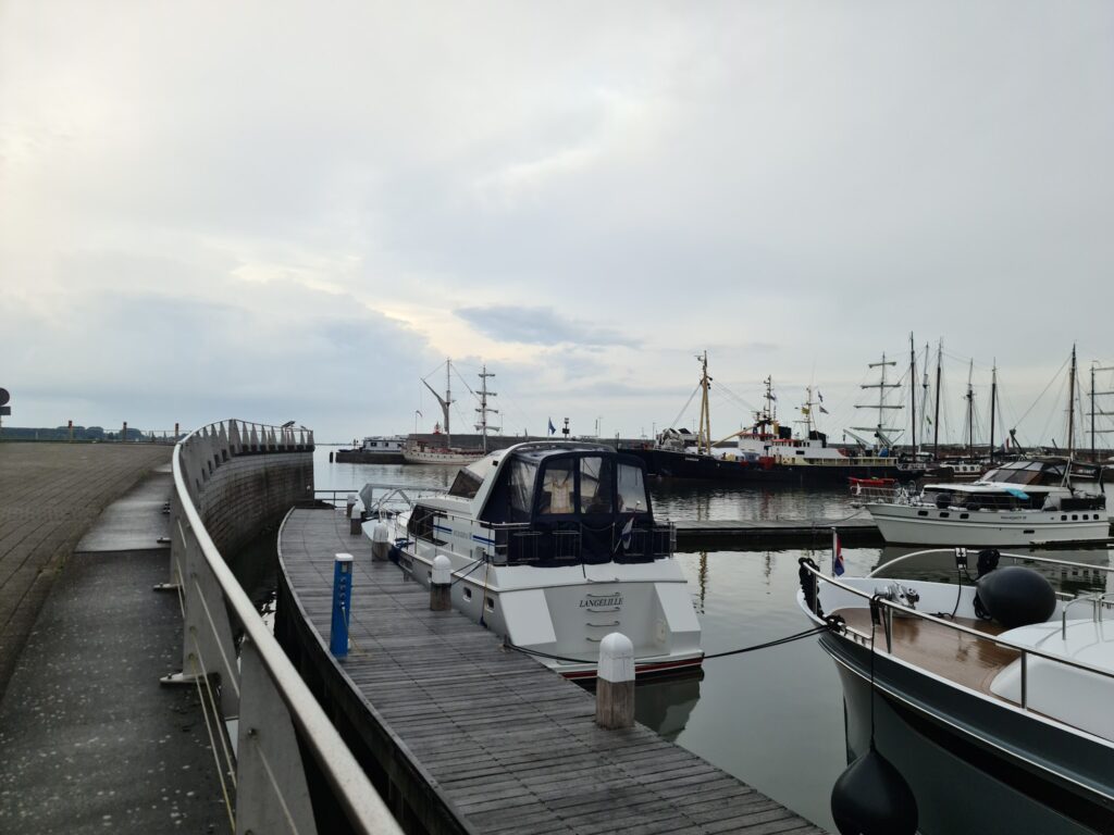 Ruhiger Hafen bei bedecktem Himmel; Motor- und Segelboote liegen an Holzstegen, Steinmole links im Bild.