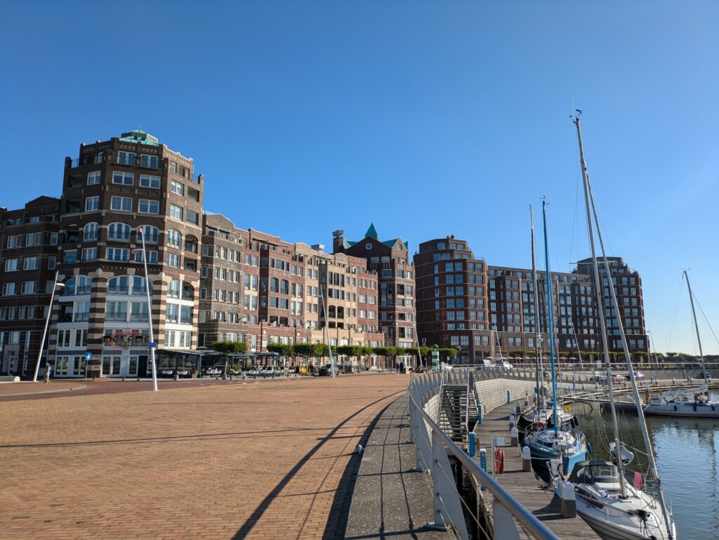 Sonnige Hafenpromenade mit Backsteinbauten; festgemachte Segelboote spiegeln sich im ruhigen Wasser.
