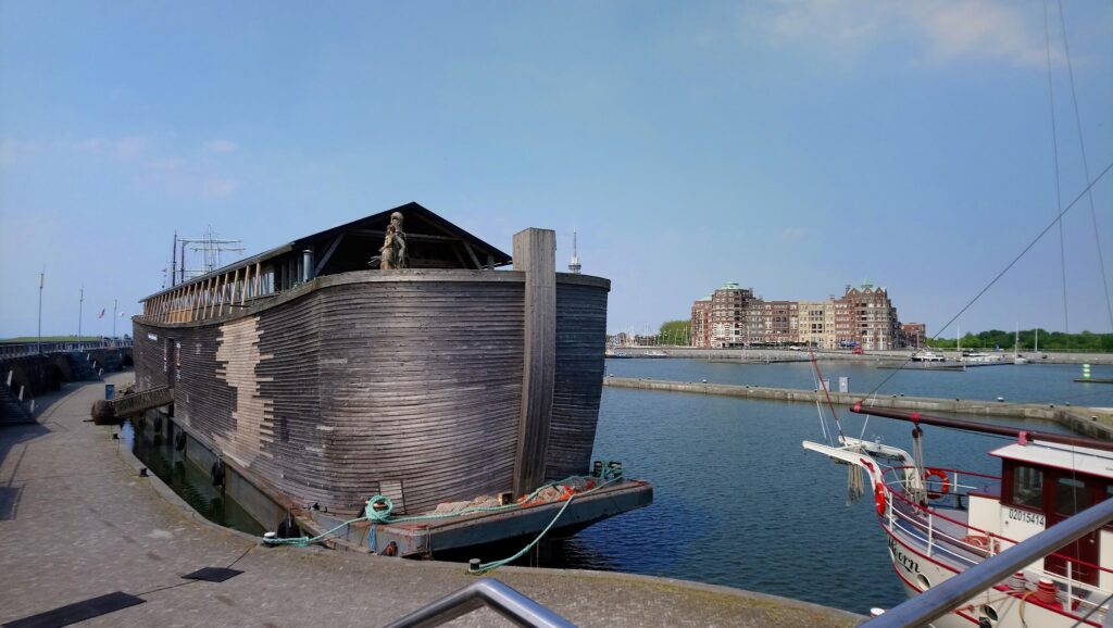 Große hölzerne Arche-Nachbildung vertäut im Hafen; rechts Segelboote, hinten Backsteinwohnblöcke unter blauem Himmel