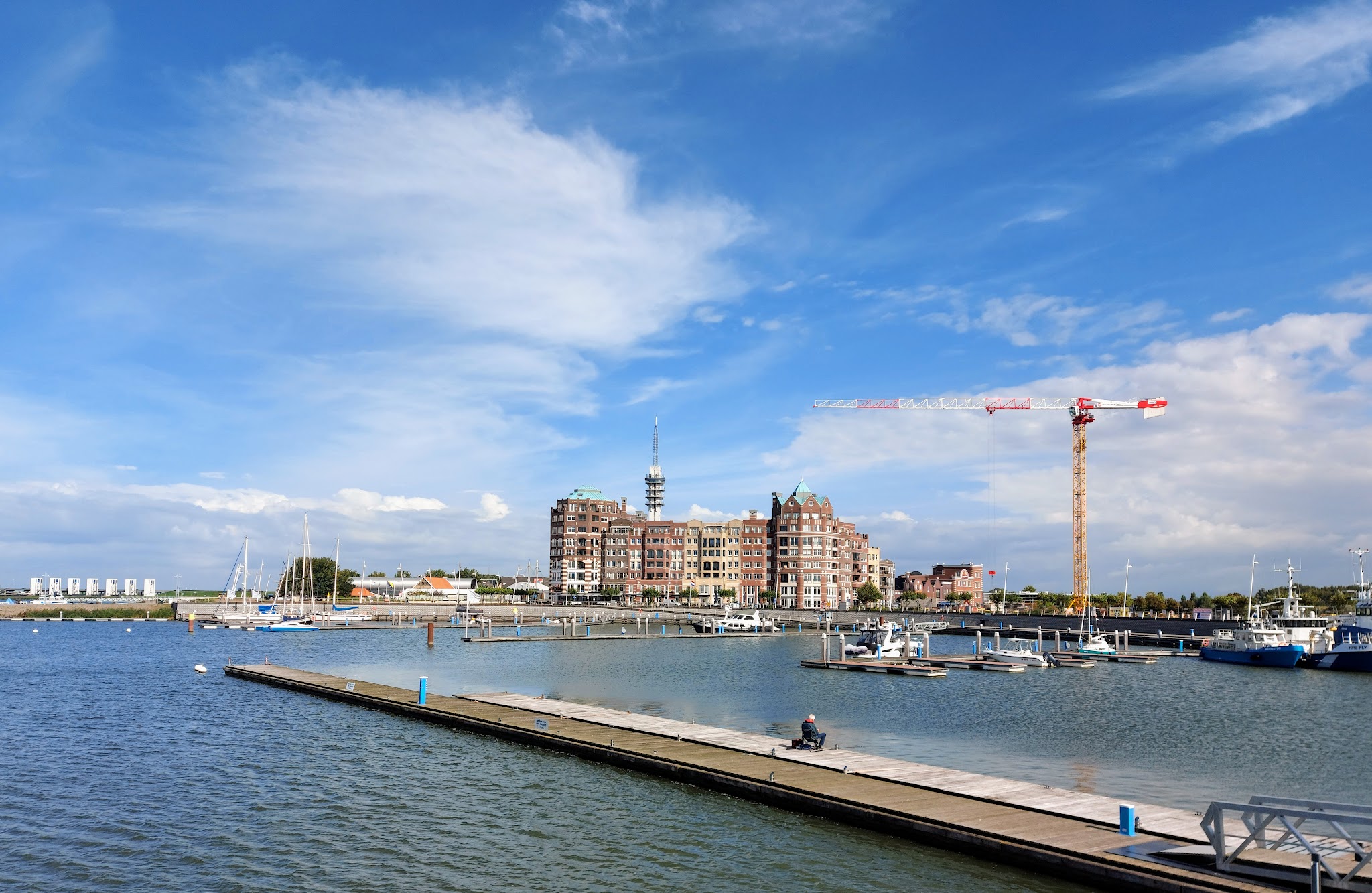 Marina mit Segelbooten und Steg, Backsteingebäude dahinter; Funkmast und Baukran unter blauem Himmel.