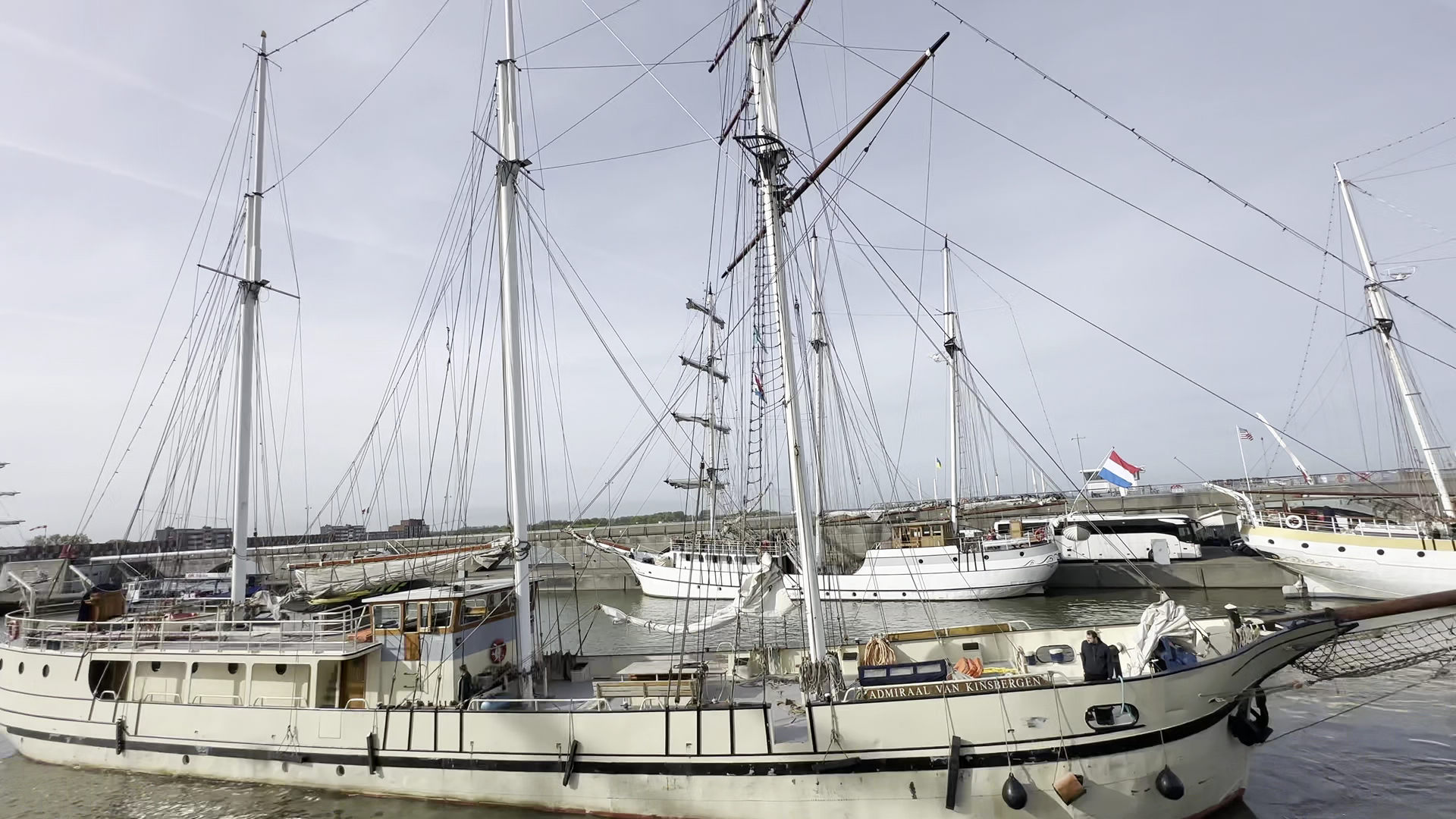 Mehrere historische Segelschiffe mit hohen Masten liegen im Hafen, eine niederländische Flagge weht.
