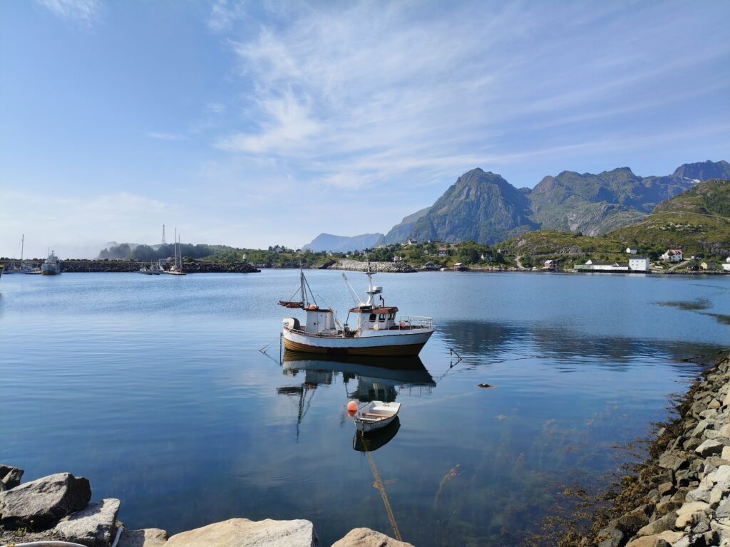 Kleines Fischerboot auf ruhigem Fjord; Berge und klarer Himmel spiegeln sich im blauen Wasser.