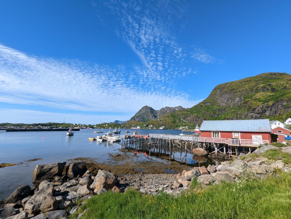 Rotes Bootshaus auf Stelzen am ruhigen Fjord; kleine Boote im Wasser, grüne Berge und blauer Himmel.