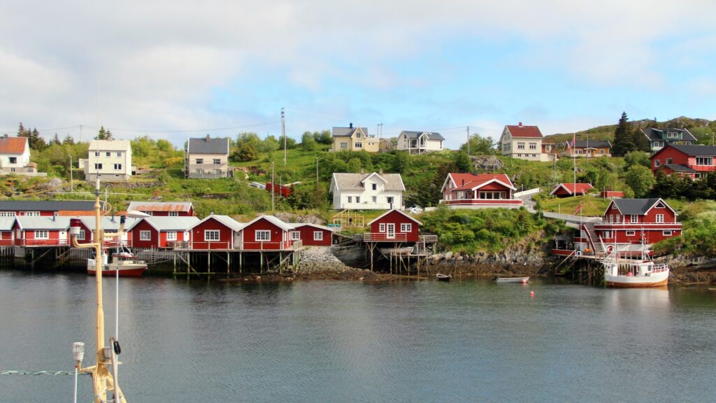 Kleine norwegische Küstensiedlung: rote Fischerhütten auf Stelzen am Wasser, Boote davor, bunte Häuser am grünen Hang
