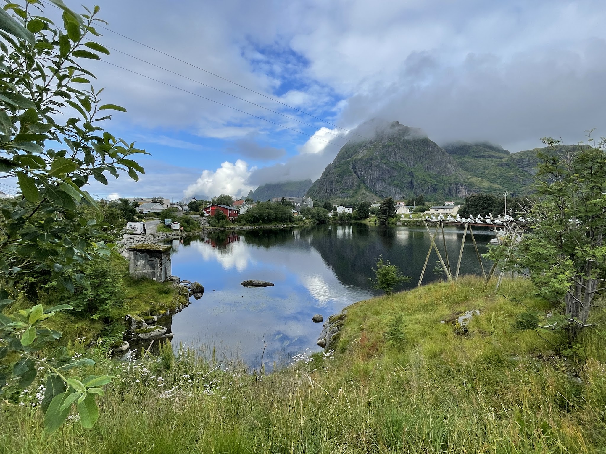 Spiegelnder See vor wolkenverhangenem Berg; rote Hütte, kleine Häuser und Möwen auf Holzgestell in grüner Landschaft.