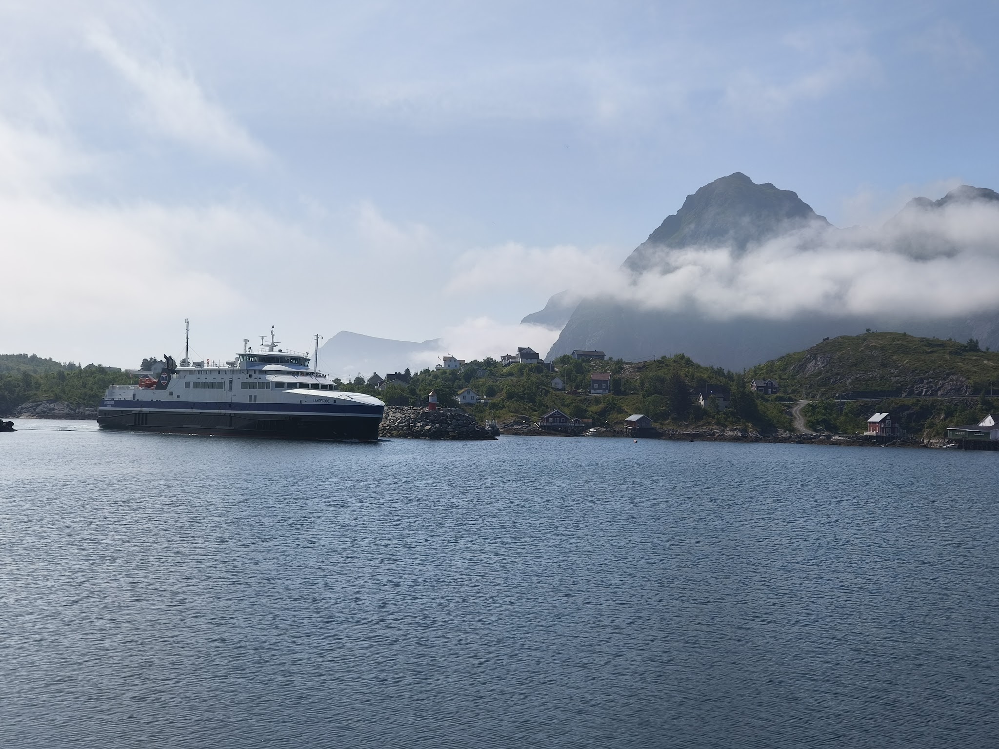 Große Fähre fährt durch ruhigen Fjord, dahinter kleines Dorf am Ufer und wolkenverhangener Berg
