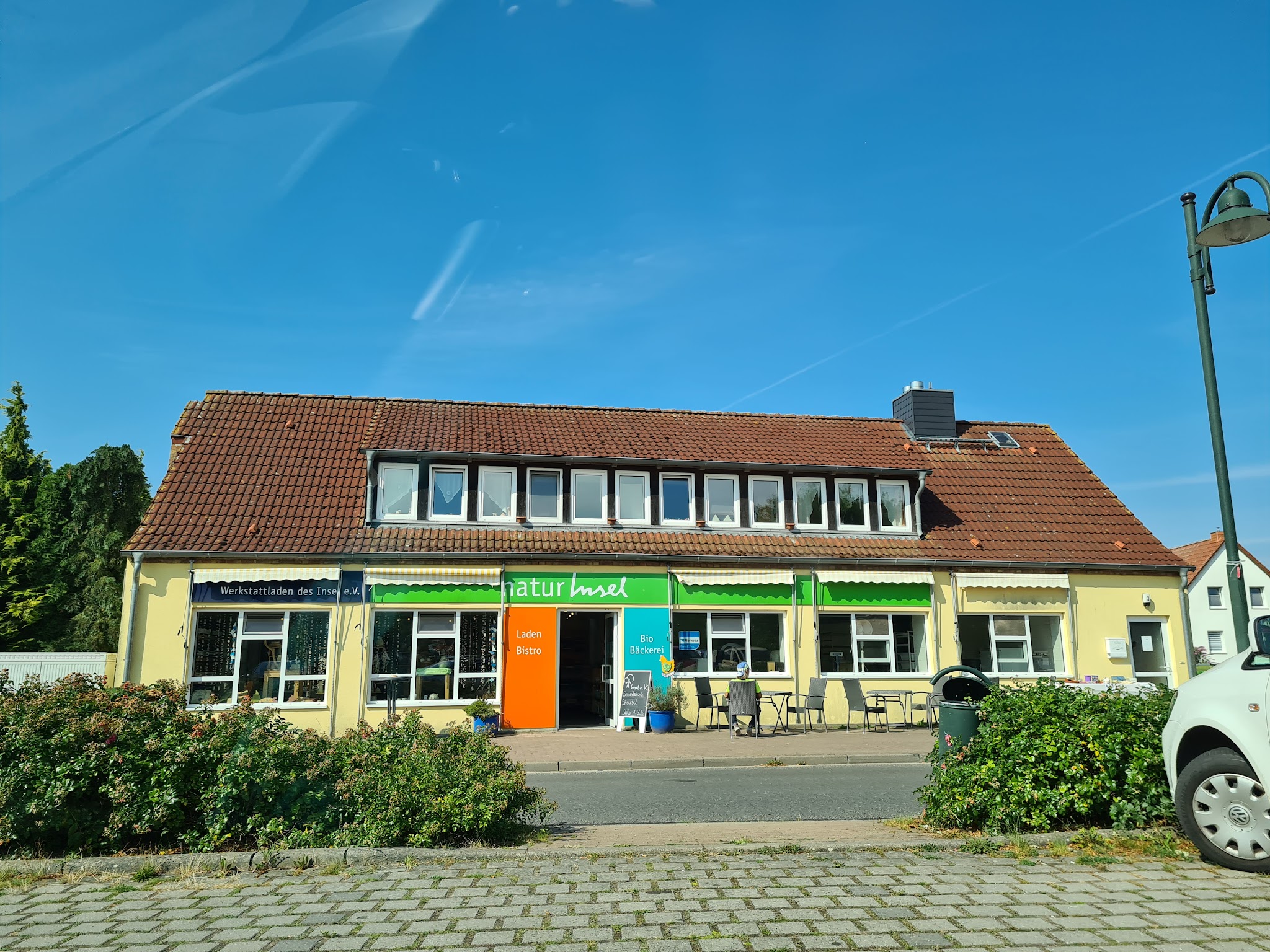 Gelbes, einstöckiges Ladencafé „naturInsel“ mit grünen Markisen und Außentischen unter blauem Himmel