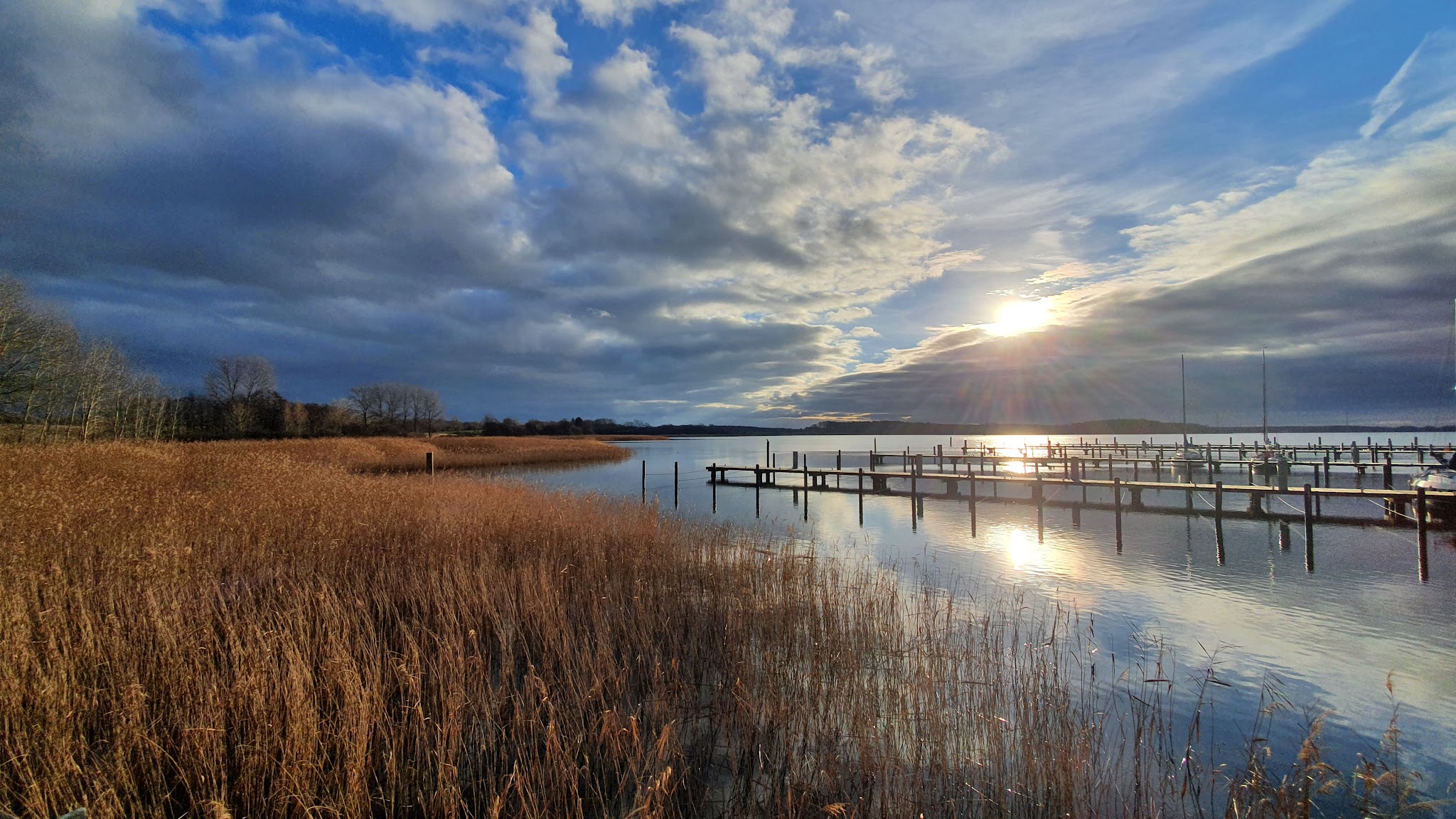Hölzerner Steg mit vertäuten Segelbooten im ruhigen See, Sonnenuntergang über Schilfufer und dramatischen Wolken.