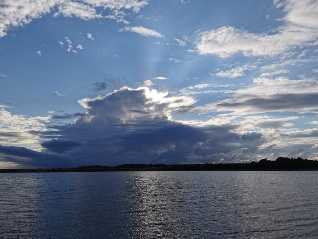 Dramatischer Himmel über ruhigem See; Sonne hinter massiver Wolke streut Strahlen, Reflexe im Wasser.