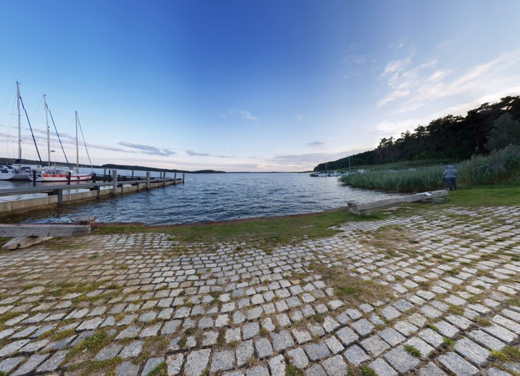 Kopfsteinpflasterufer mit Steg und Segelbooten an ruhigem See, Person steht rechts an Schilf vor Wald.