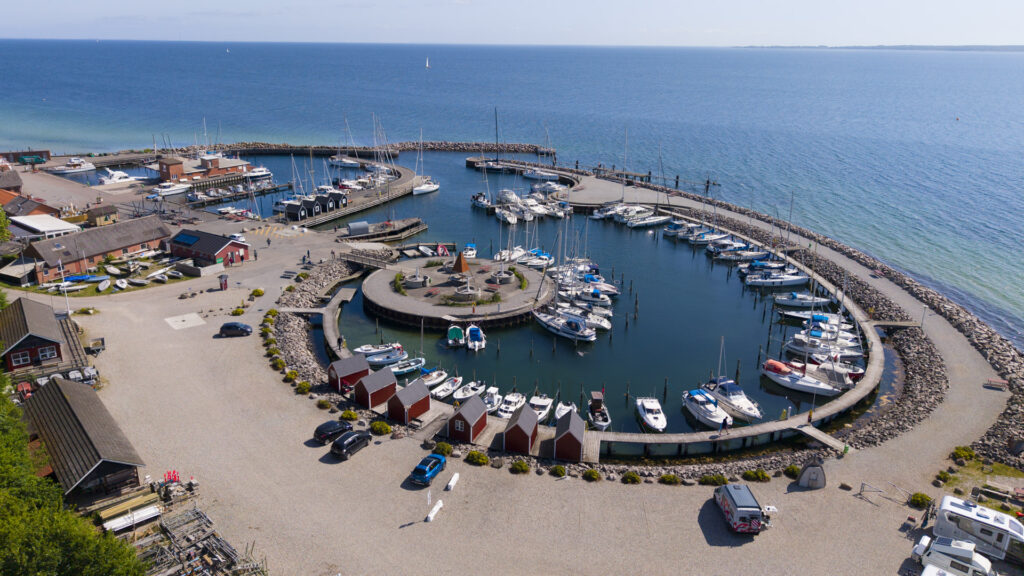 Luftaufnahme eines kreisförmig angelegten Yachthafens mit vielen Segelbooten, roten Bootshäuschen und offenem Meer.