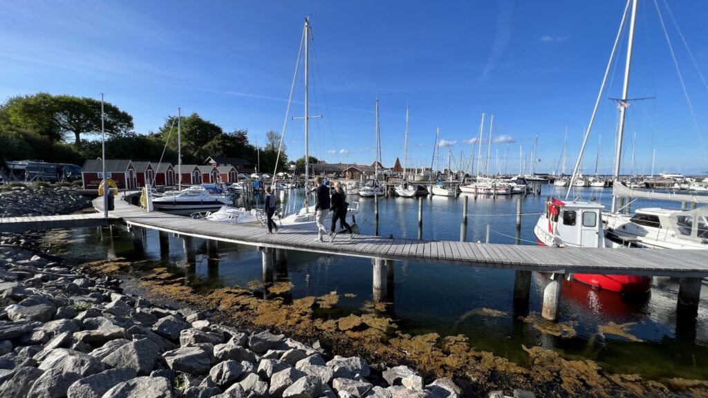 Mehrere Segelboote im kleinen Hafen; drei Personen gehen über Steg, rote Bootsschuppen und blauer Himmel.