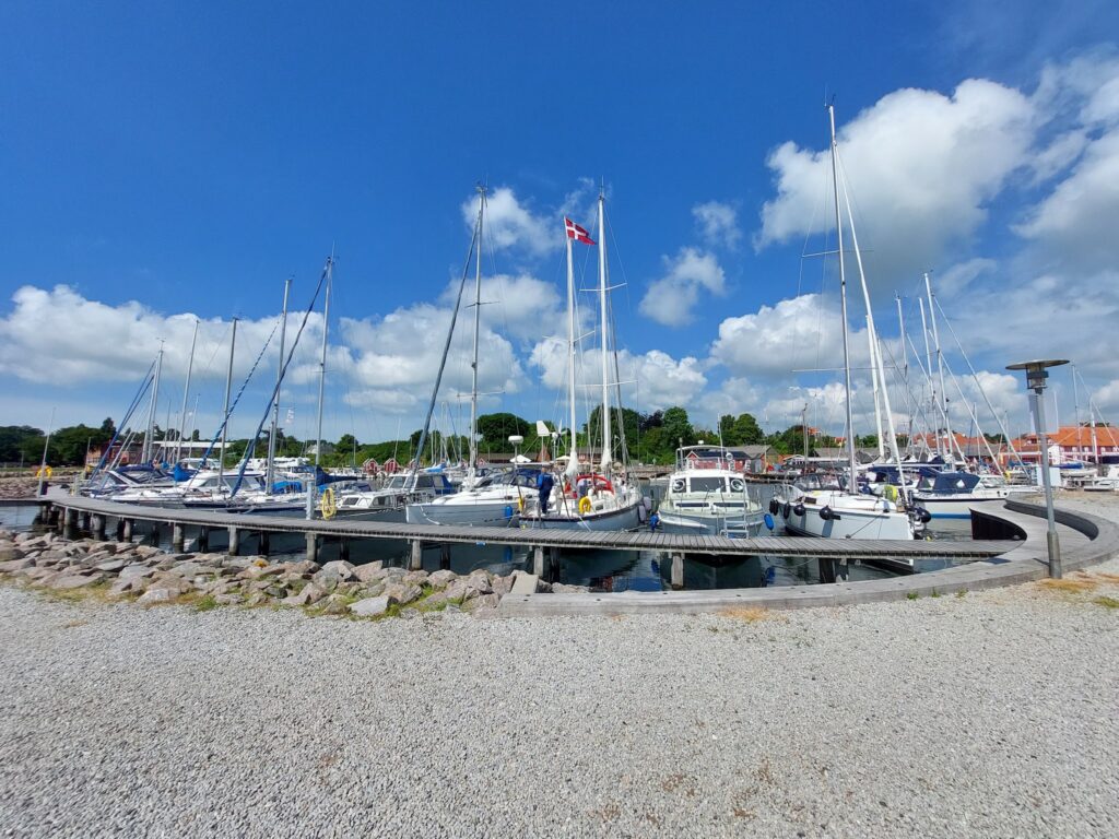Mehrere Segelboote liegen an einem Holzsteg im Hafen; über ihnen weht bei blauem Himmel eine dänische Flagge.