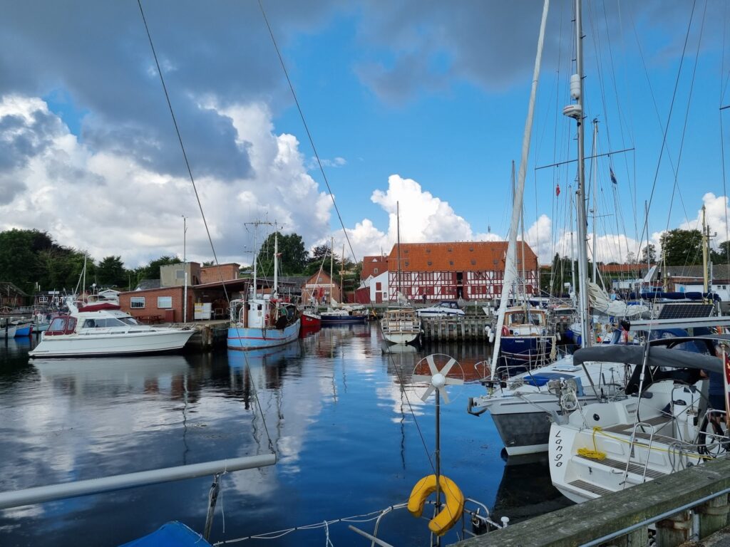 Kleiner Hafen mit Segel- und Fischerbooten; rotes Fachwerkhaus am Kai, Wasser spiegelt Wolken im blauen Himmel.