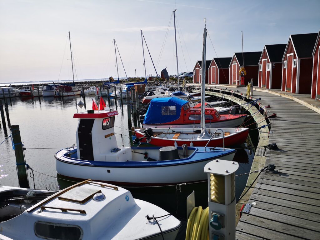 Ruhiger Hafen mit kleinen Segel- und Motorbooten an Holzsteg, Reihe roter Bootshäuser im Hintergrund.
