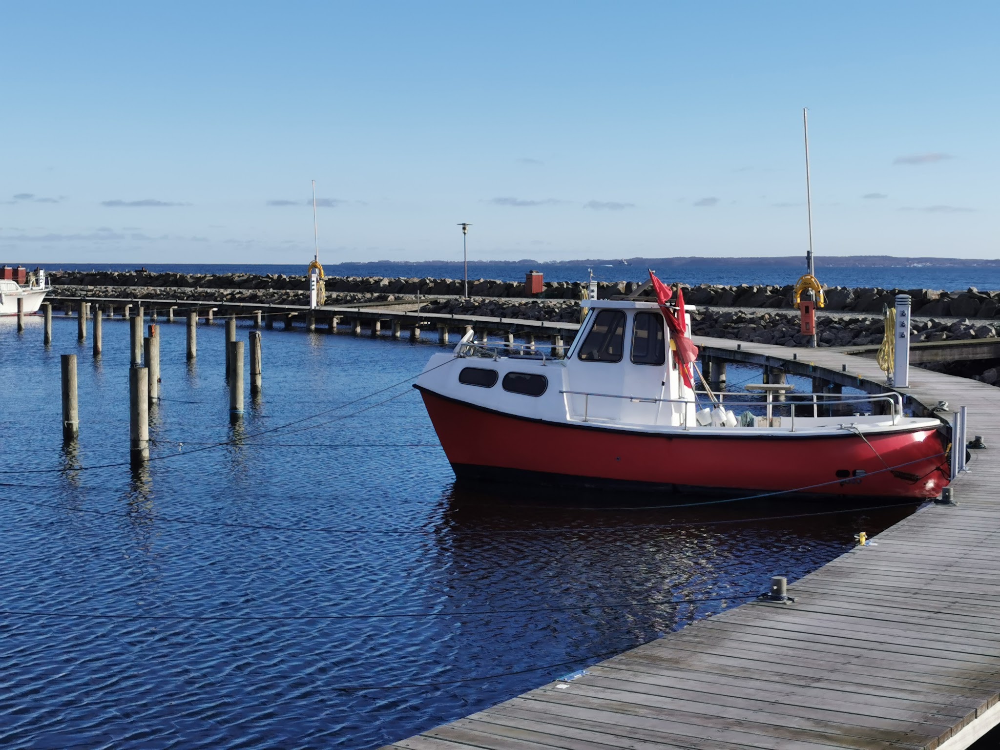 Kleines rot-weißes Boot mit roter Flagge liegt an Holzsteg in ruhigem Hafen unter klarem blauem Himmel.