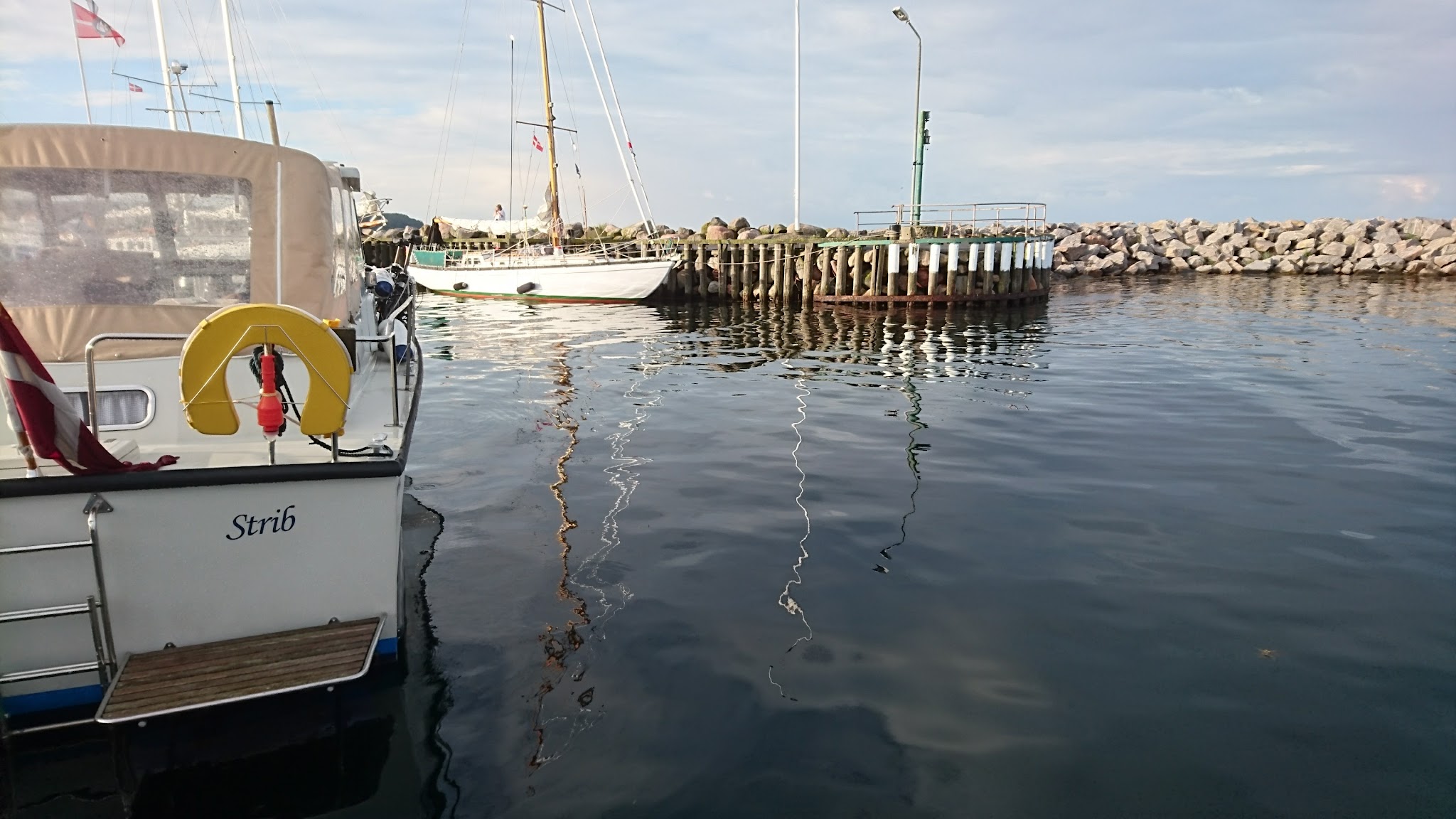 Segelboot im ruhigen Hafen; gelber Lebensring am Heck, Masten spiegeln sich im glatten Wasser vor Steinmole.