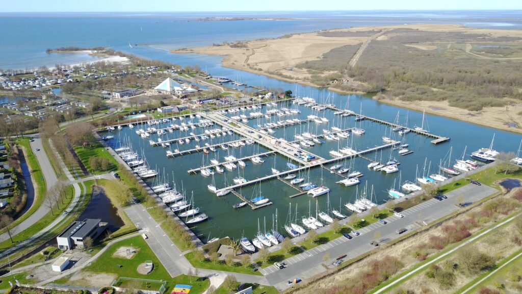 Luftaufnahme eines Yachthafens voller Segelboote, von schmalen Stegen umgeben, dahinter Küste und weite See.
