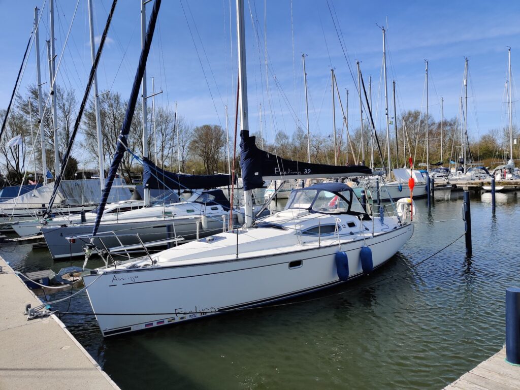 Weißes Segelboot im Yachthafen, umgeben von vielen Masten; ruhiges Wasser unter klarem, blauem Himmel.