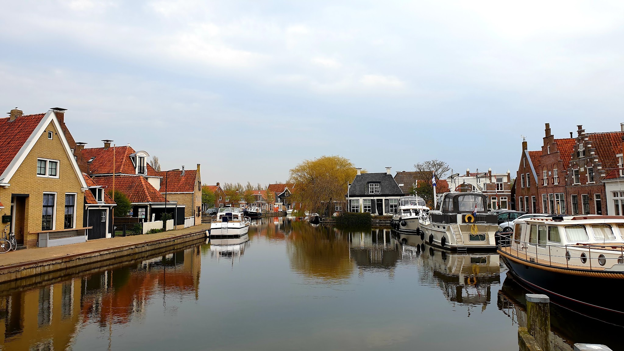 Ruhiger Kanal mit Booten, flankiert von roten Ziegelhäusern; Spiegelung im Wasser unter bewölktem Himmel