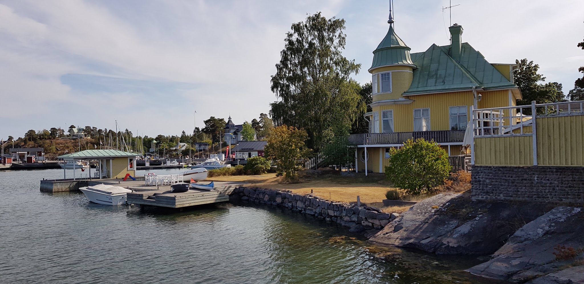Gelbes Holzhaus mit grünem Turmdach am felsigen Ufer; Steg mit kleinen Motorbooten in ruhigem Inselhafen.