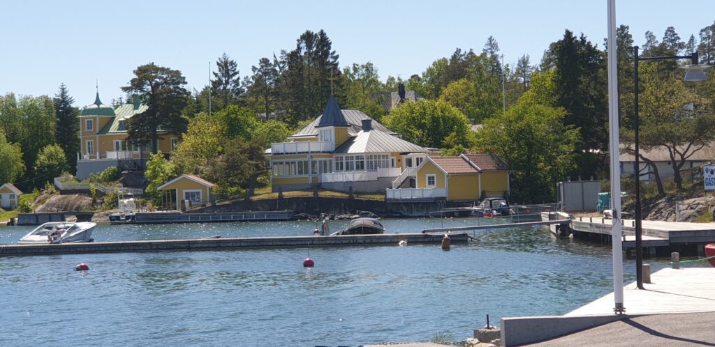 Gelbe Holzvillen zwischen Bäumen am schwedischen Ufer; davor Anleger mit Motorbooten im klaren Wasser.