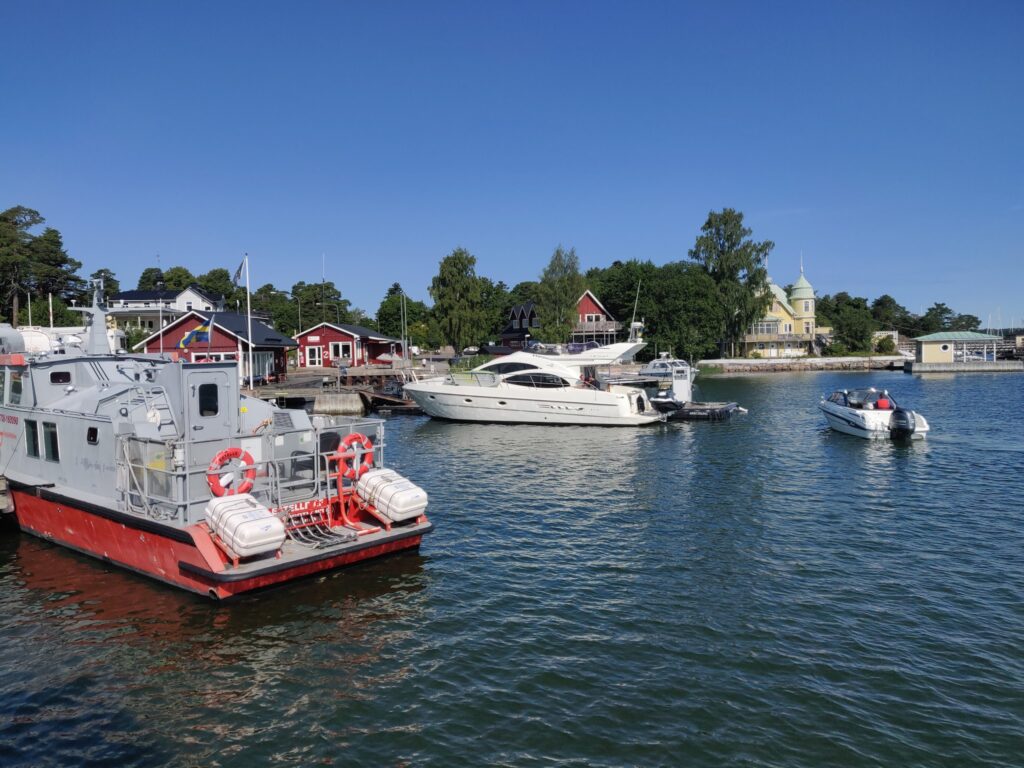 Kleiner Hafen bei klarem Himmel: Rettungsboot, Yacht und Motorboot vor roten Holzhäusern und grünem Ufer.