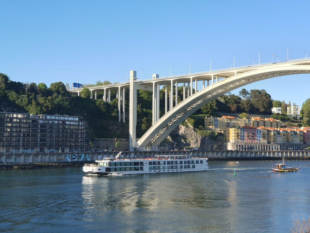 Flusskreuzfahrtschiff unter heller Bogenbrücke, dahinter bunte Häuser am bewaldeten Hang unter klarem Himmel.