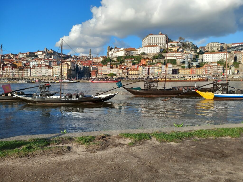 Drei Rabelo-Boote mit Weinfässern treiben vor Portos bunter Uferkulisse am Douro unter blauem Himmel mit Wolken