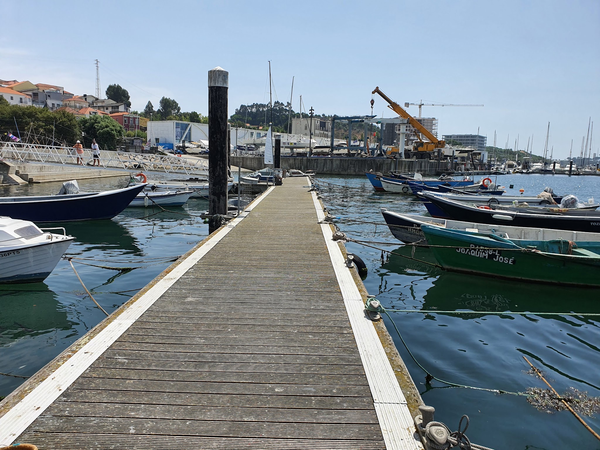 Holzsteg führt zwischen vertäuten kleinen Booten ins Hafenbecken; ruhiges Wasser, Häuser und Kran im Hintergrund.