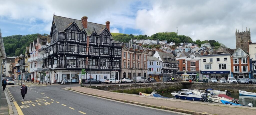 Historische Fachwerk- und viktorianische Häuser entlang Hafenstraße; kleine Boote im Wasser, grüner Hügel im Hintergrund