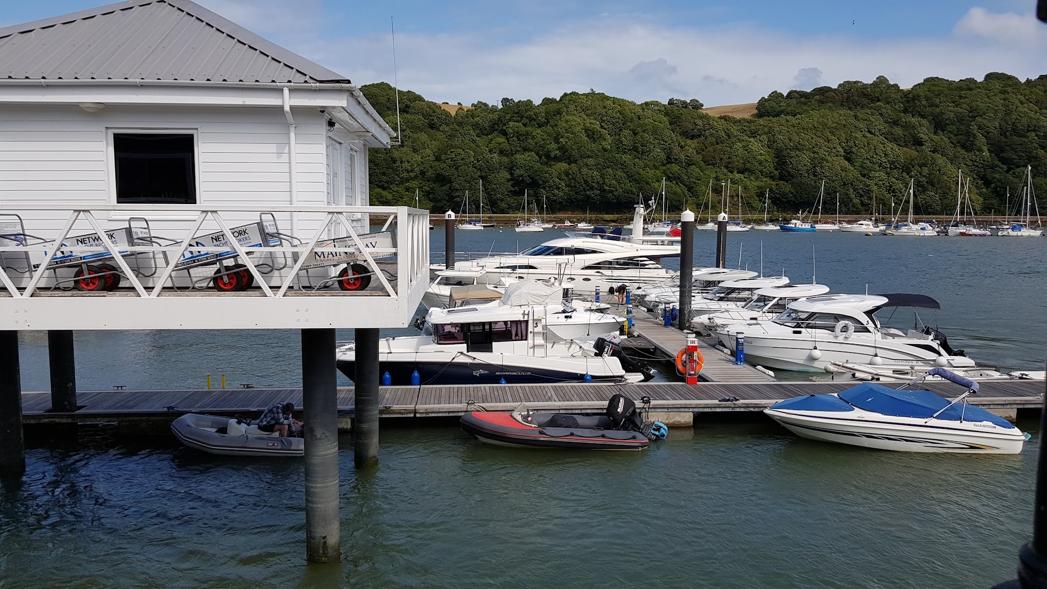 Bootssteg mit mehreren weißen Motoryachten, links ein Pfahlhaus auf Stelzen; grüne Hügel im Hintergrund.