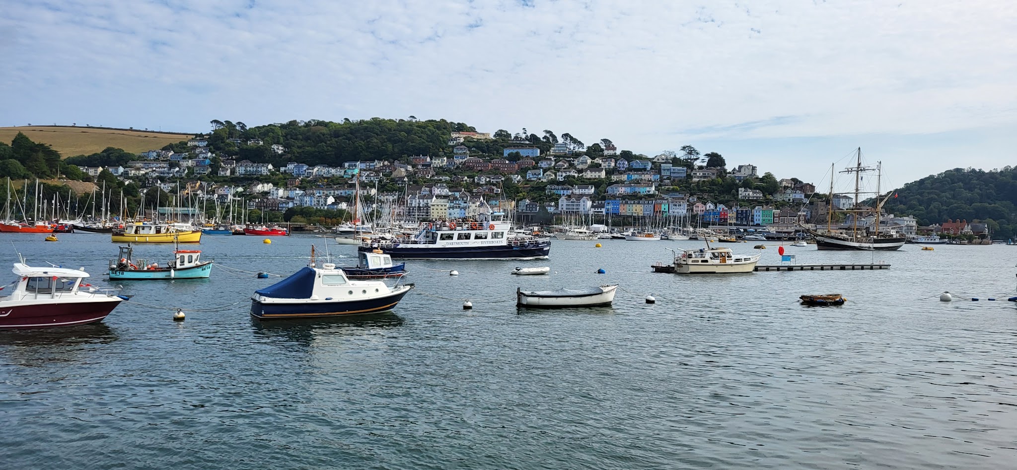 Bunte Küstenstadt am Hang, Segelboote und Schiffe ankern im ruhigen Hafen unter leicht bewölktem Himmel.