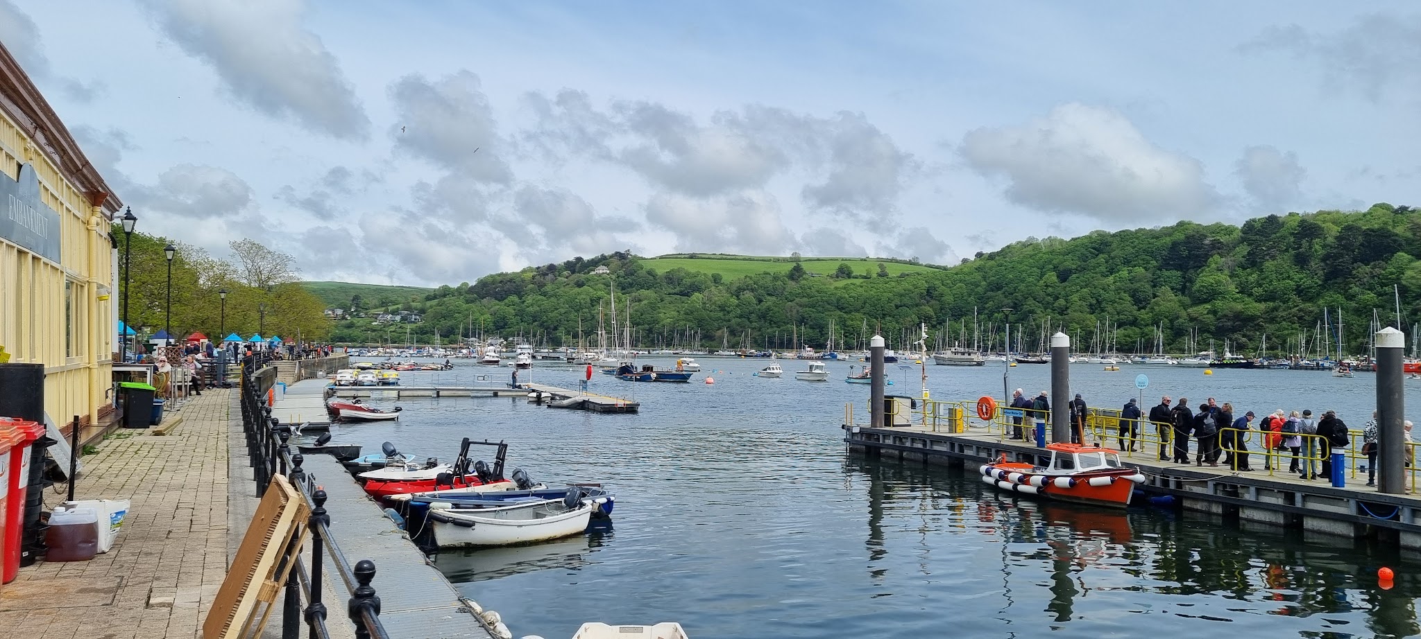 Belebter Hafen: Menschen spazieren am Kai, bunte Boote liegen im Wasser vor grünem Hügel unter bewölktem Himmel
