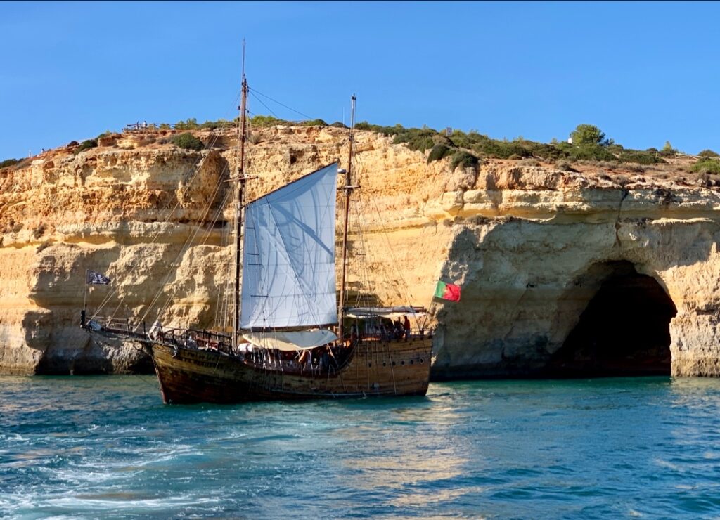 Historischer Holzsegler mit weißem Hauptsegel und portugiesischer Flagge vor hellen Felsklippen und Meeresgrotte