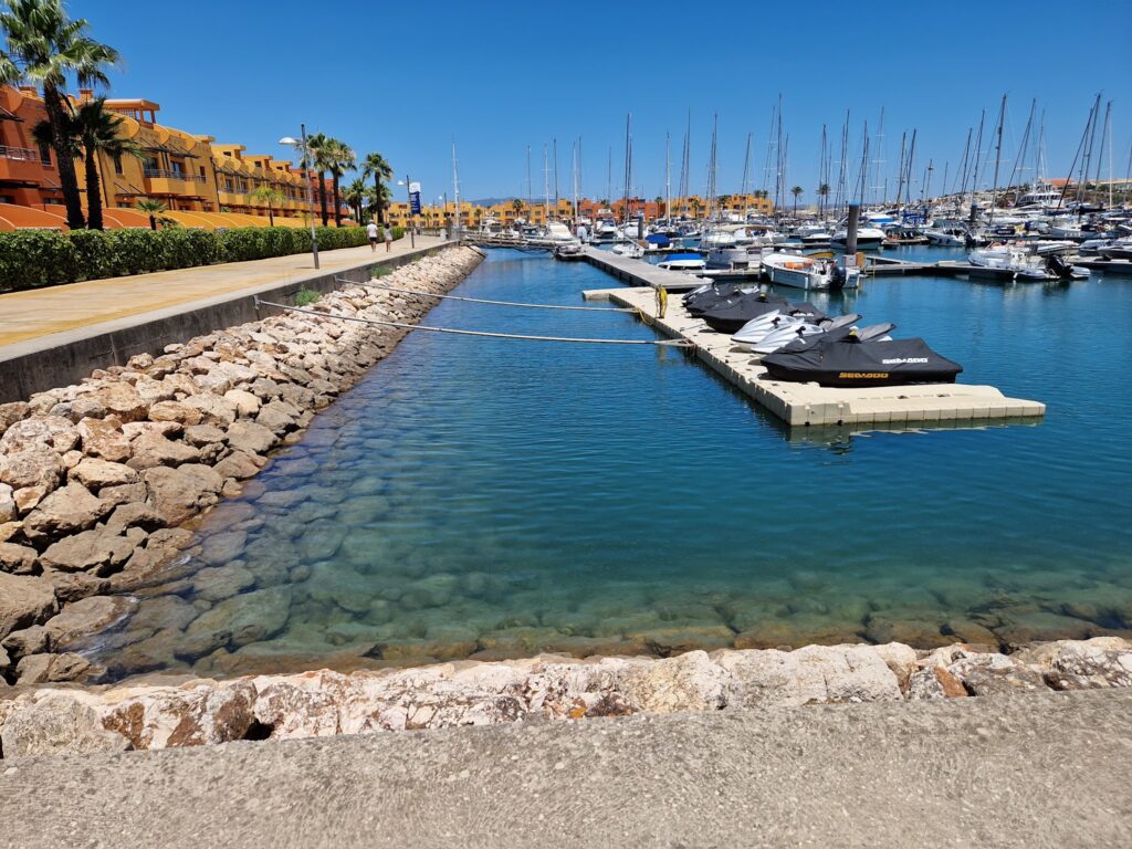 Jachthafen mit kristallblauem Wasser, Jet-Skis auf Steg, dahinter Segelboote, Promenade mit Palmen und orange Häusern.
