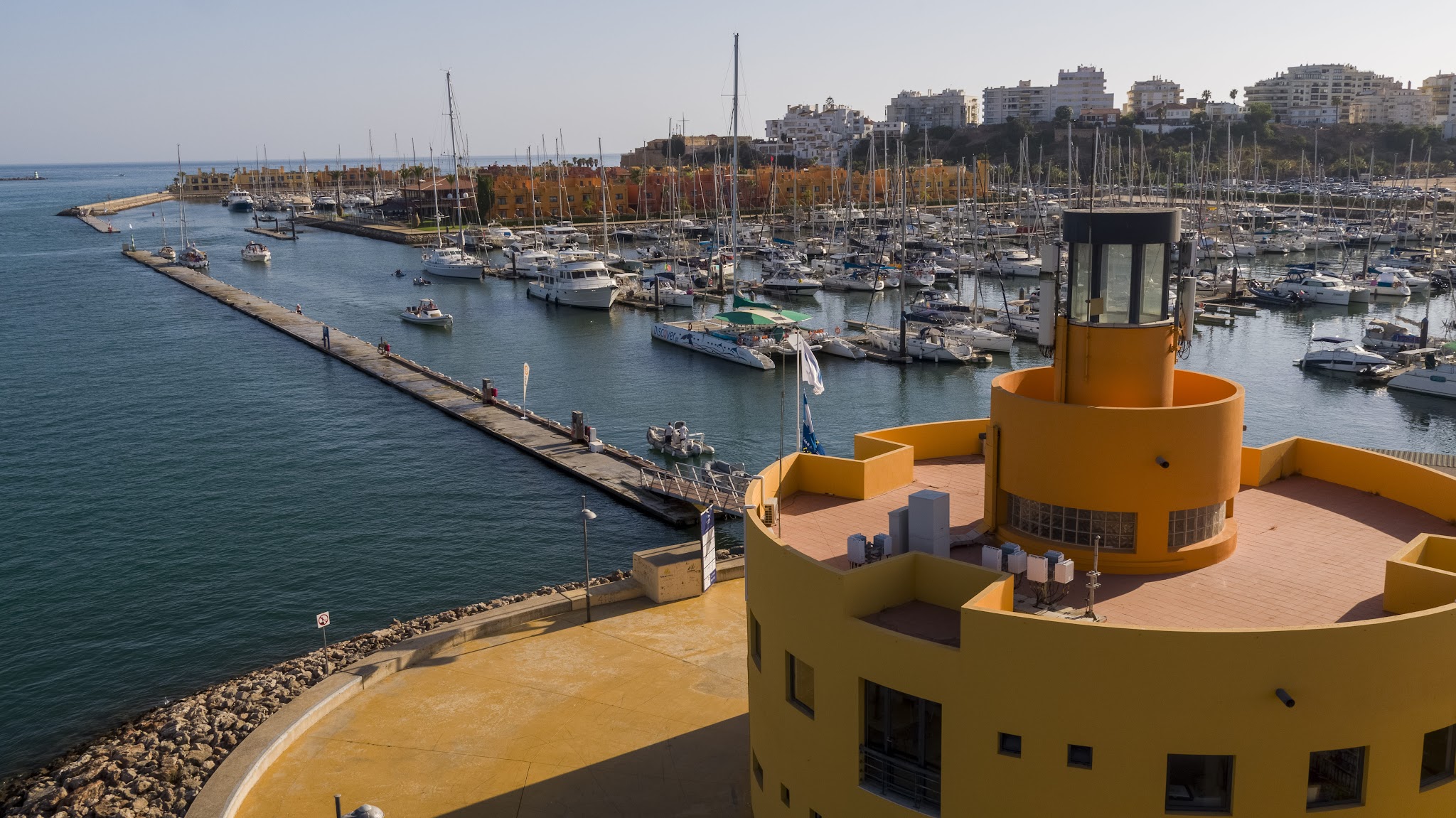 Gelber runder Leuchtturm am Hafenbecken, davor Steg, dahinter zahlreiche Segel- und Motorboote im Yachthafen.