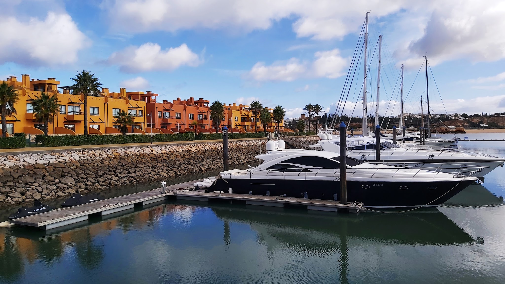 Mehrere Yachten im ruhigen Hafen, dahinter bunte mediterrane Häuser und Palmen unter leicht bewölktem Himmel