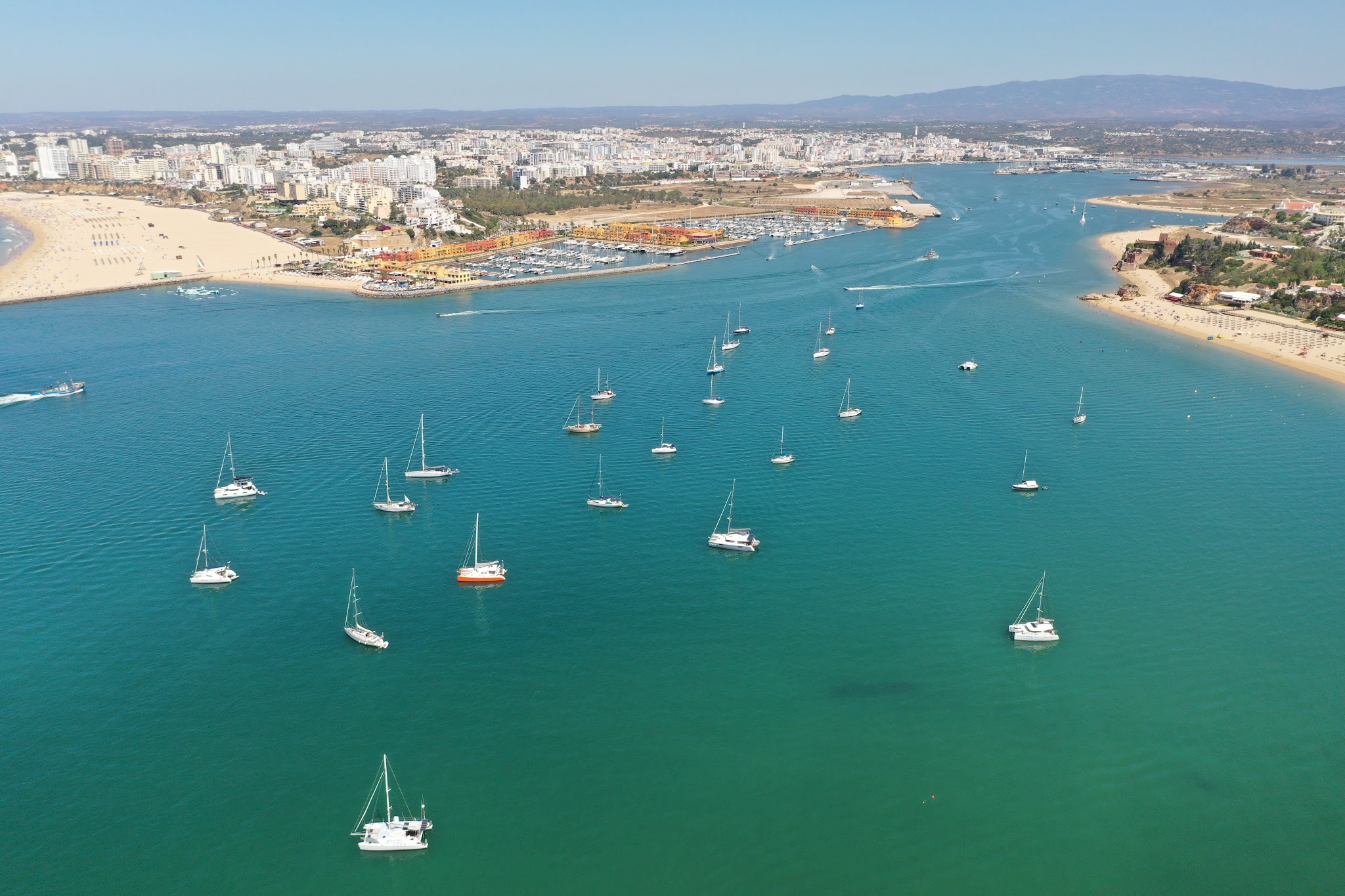 Drohnenblick auf türkisfarbenes Hafenbecken mit verstreuten Segelbooten, eingerahmt von Stränden und Stadt.