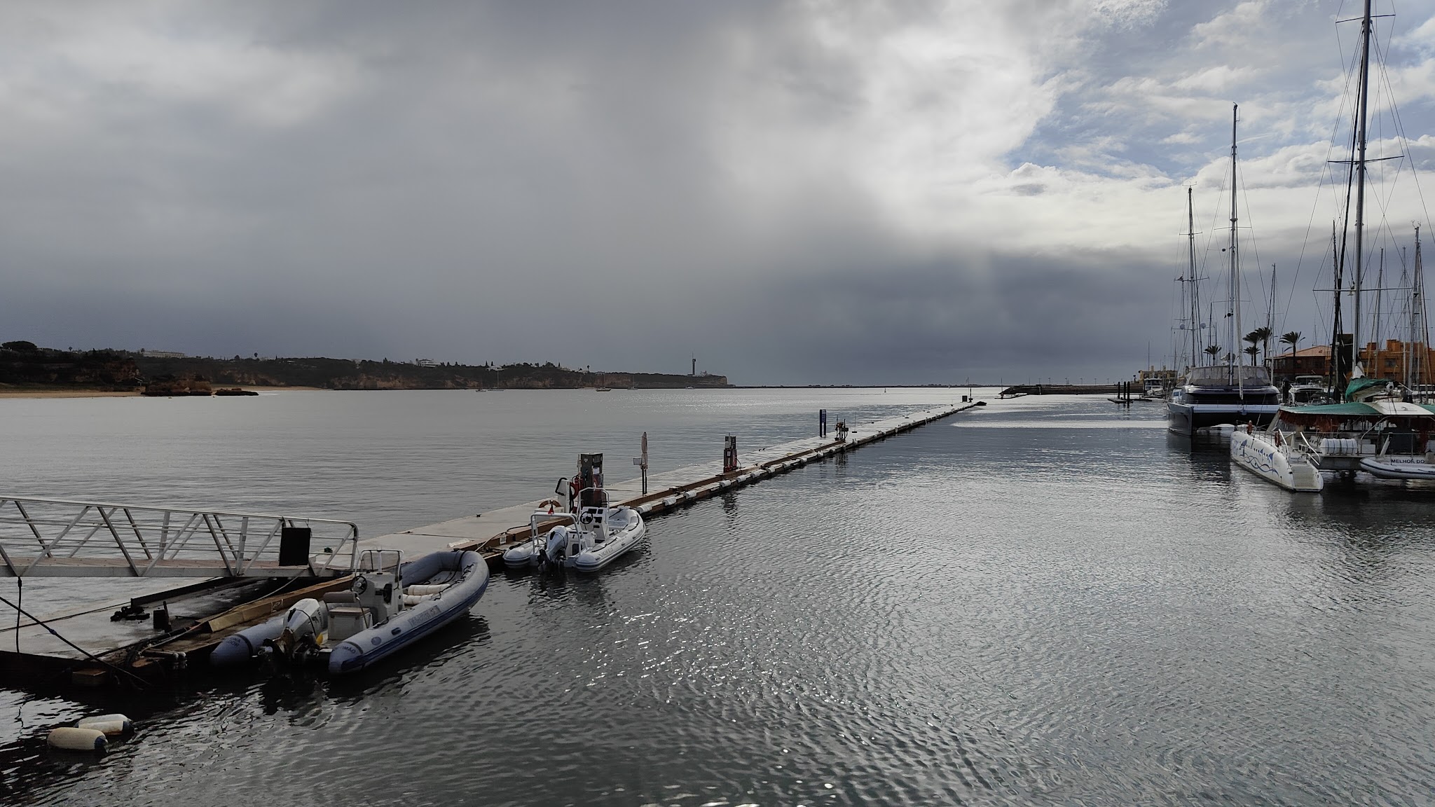 Schmale Mole mit kleinen Schlauchbooten, rechts Segelboote im Hafen; ruhiges Wasser unter dicht bewölktem Himmel.