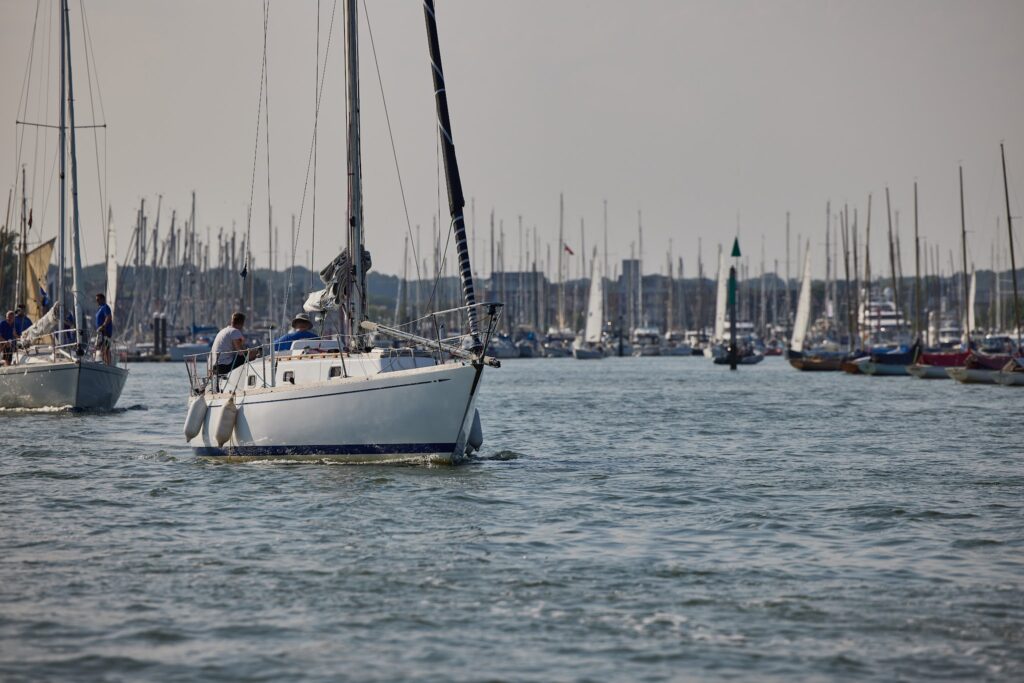 Segelboot mit Crew verlässt belebten Hafen; zahlreiche Masten weiterer Yachten im Hintergrund.