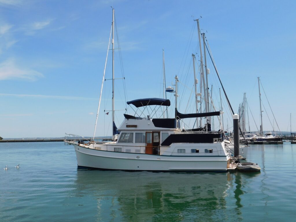 Motorjacht mit dunklem Sonnendach liegt im ruhigen Yachthafen; Segelmasten und klarer Himmel im Hintergrund.