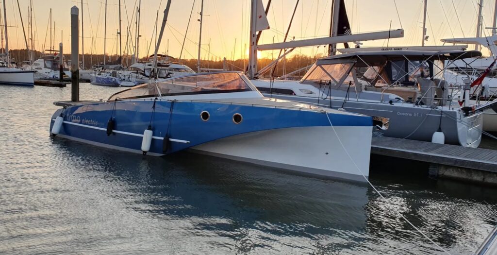 Blau-weißer Elektro-Katamaran liegt im Yachthafen; Masten im Gegenlicht eines orangefarbenen Sonnenuntergangs