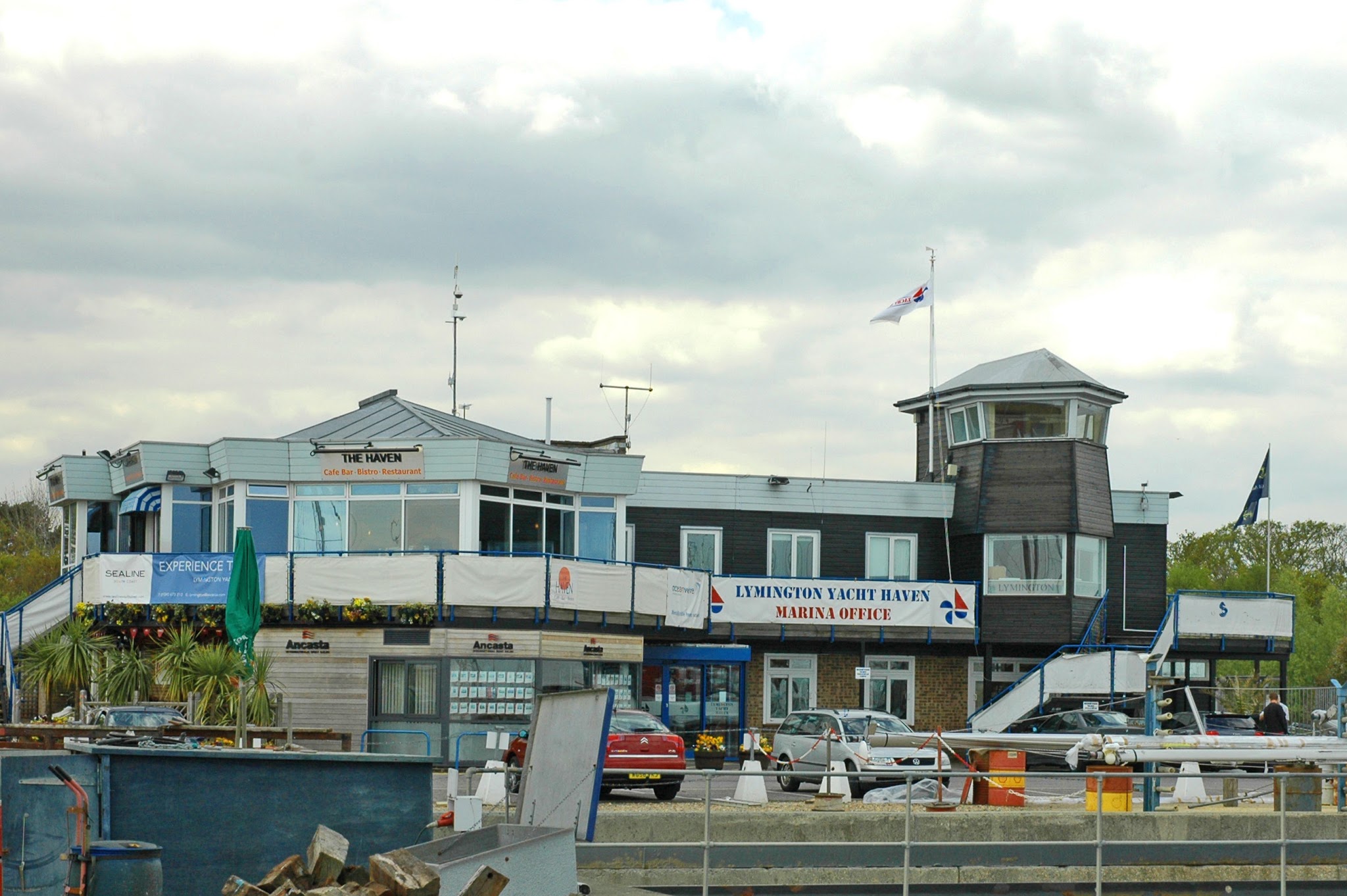 Marina-Büro mit Restaurant-Terrasse am Hafen, davor parkende Autos und Hafenanlagen unter bedecktem Himmel.