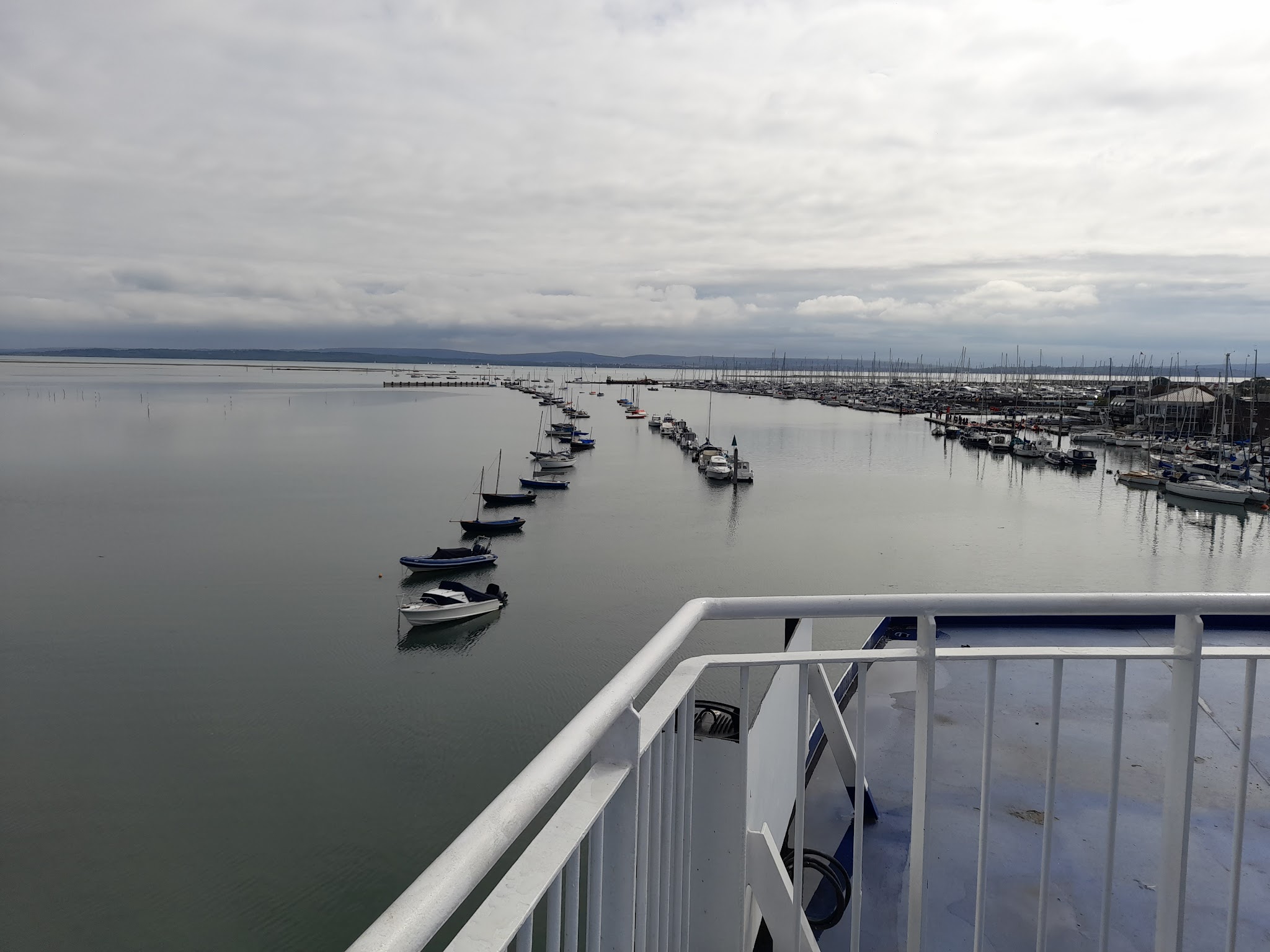 Blick von einer Fähre auf ruhigen Hafen; Boote reihen sich an Steg, hinten weitläufige Marina unter bewölktem Himmel.