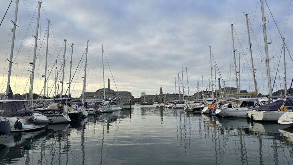 Segelboote im Hafen; schlanke Masten spiegeln sich im ruhigen Wasser vor historischen Ufergebäuden bei bedecktem Himmel.
