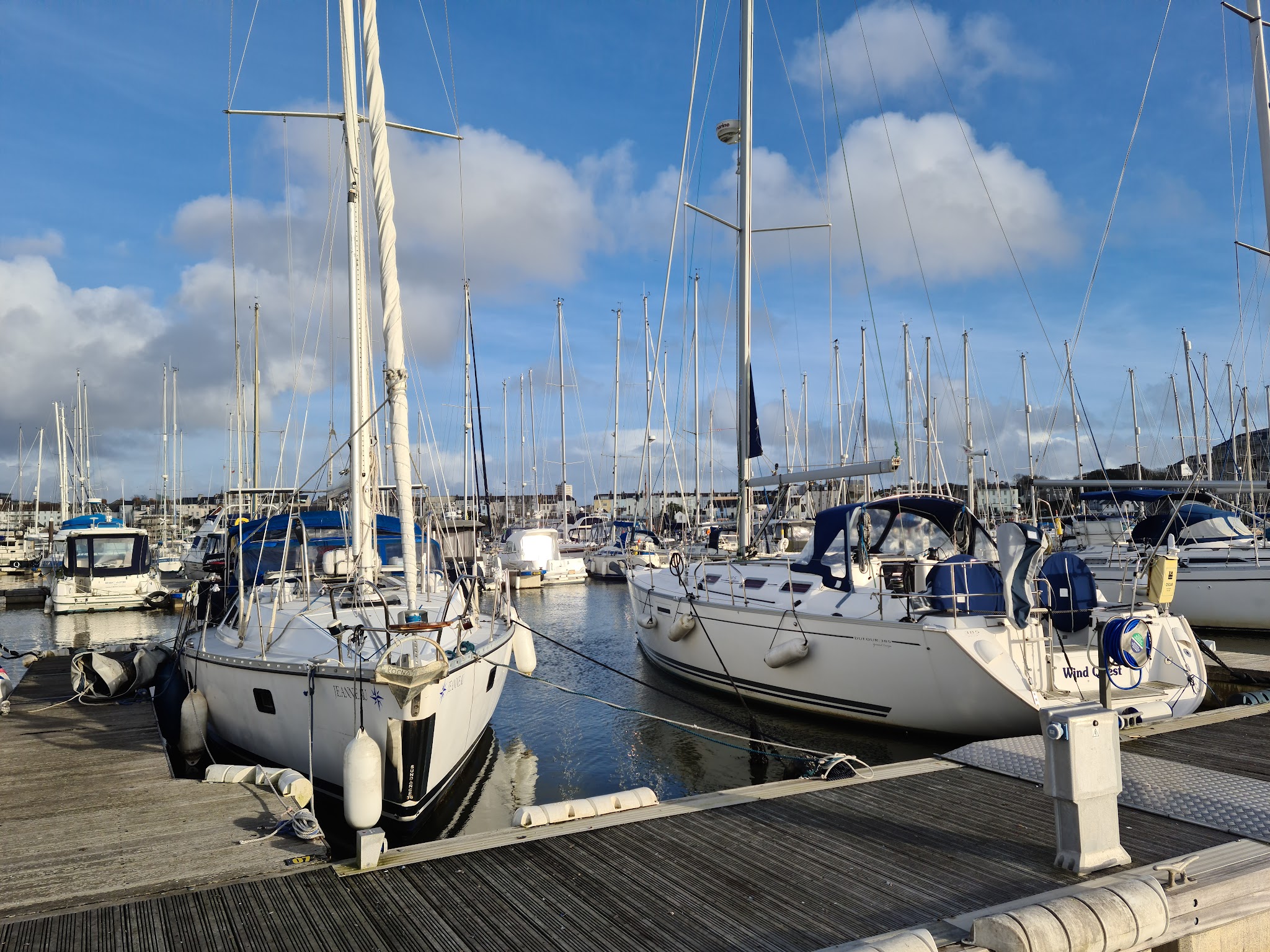 Zwei Segelboote vertäut an Stegen in ruhigem Yachthafen, zahlreiche Masten und blauer Himmel mit Wolken dahinter.