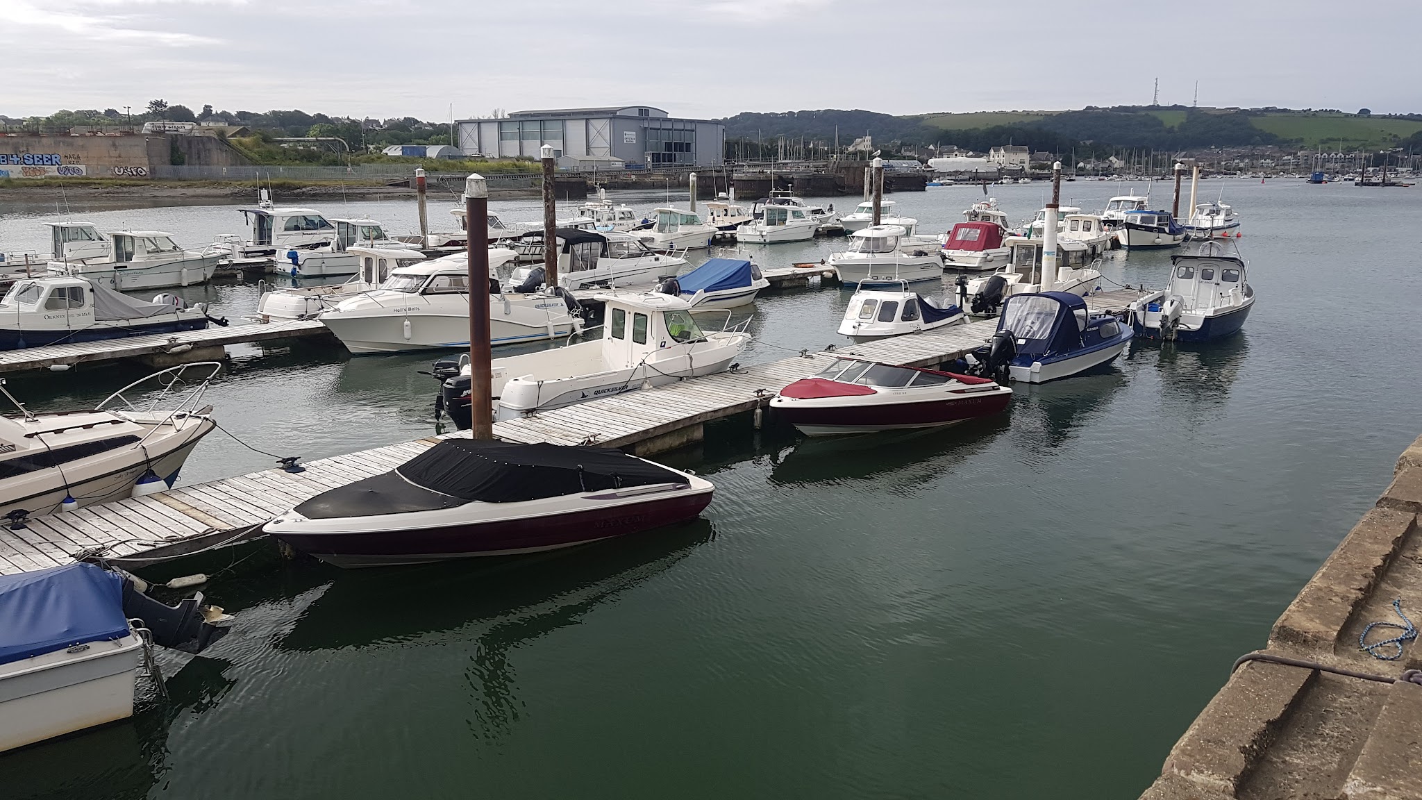 Kleine Motorboote liegen an schwimmenden Stegen im ruhigen Hafen; im Hintergrund Hangar und grüne Hügel.