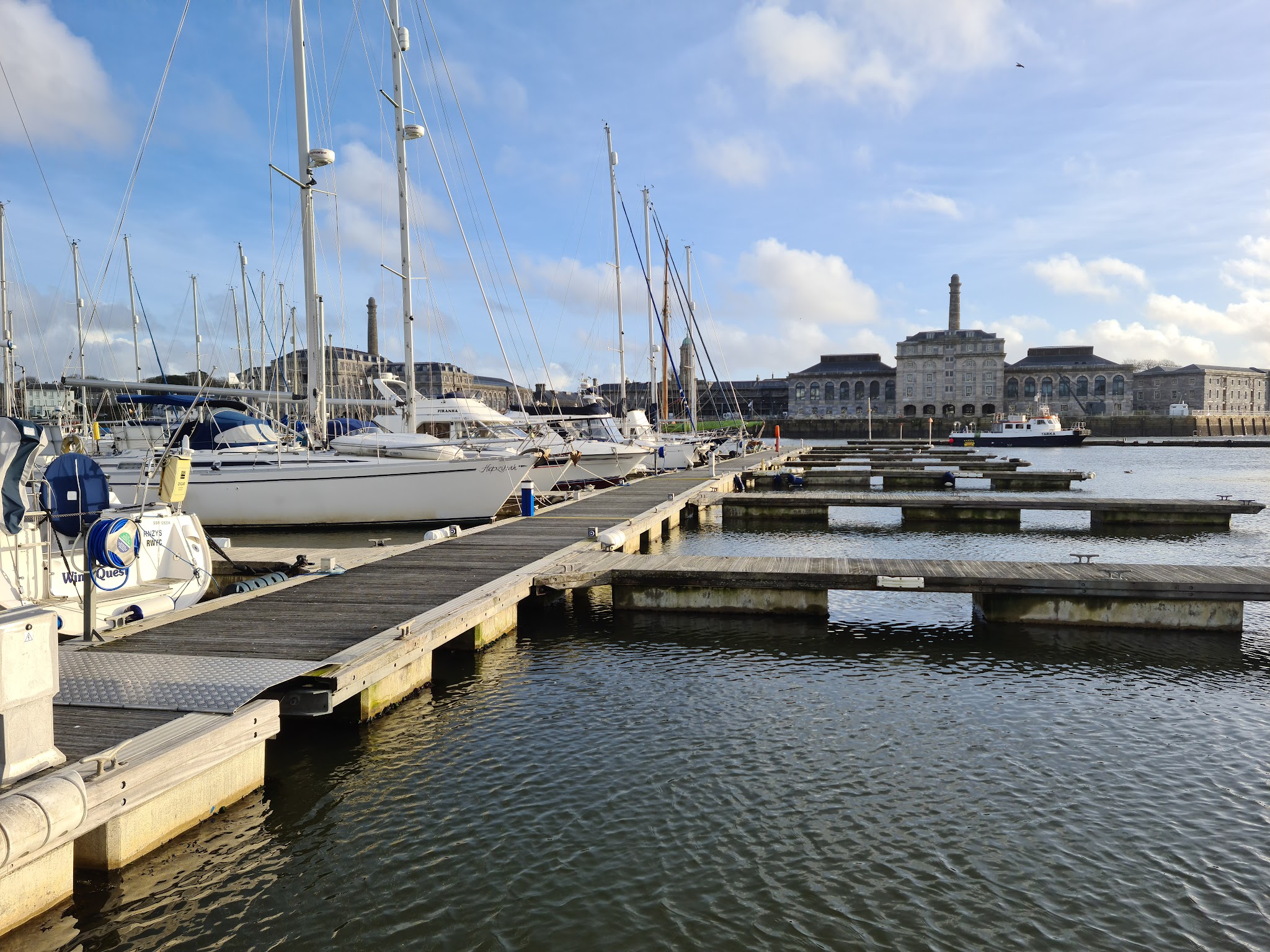 Yachthafen mit Segelbooten und leeren Stegen; dahinter großes historisches Gebäude unter blauem Himmel.