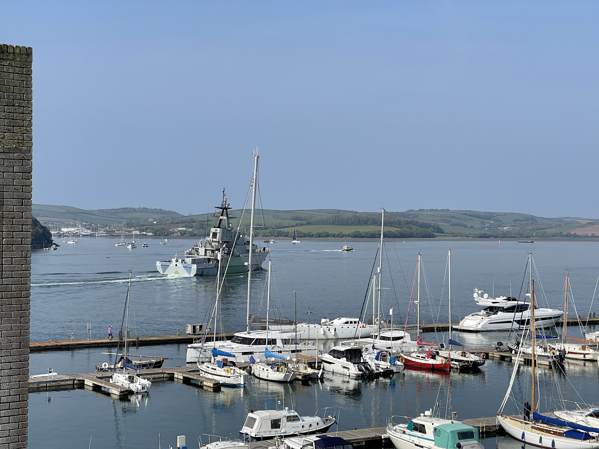 Jachthafen mit Segelbooten; graues Marineschiff passiert auf ruhigem Wasser, grüne Hügel im Hintergrund.
